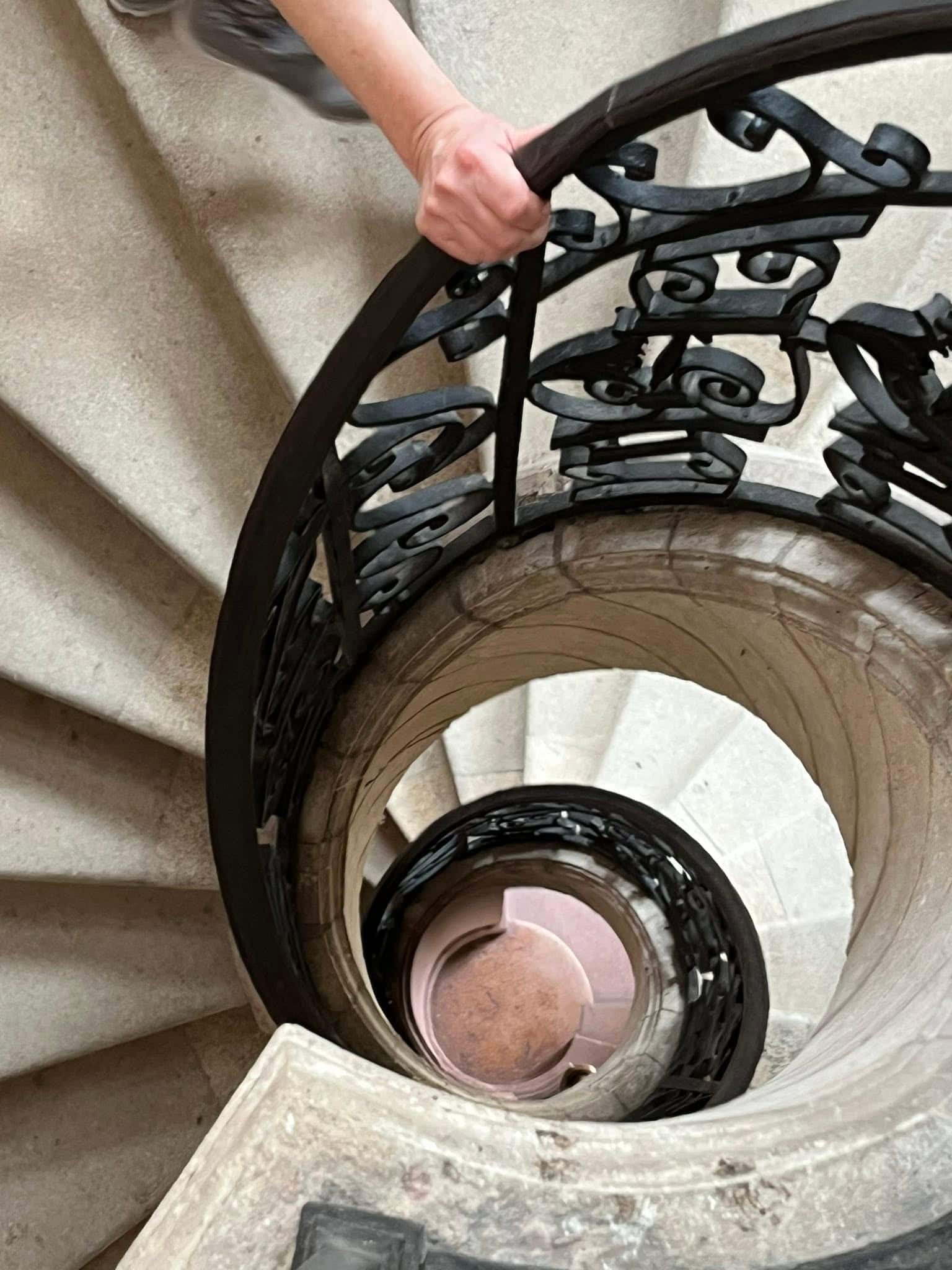 727322035_1306129295008227_901114928677831086_n Close-up of a hand gripping a black wrought-iron spiral staircase railing on a stone staircase.