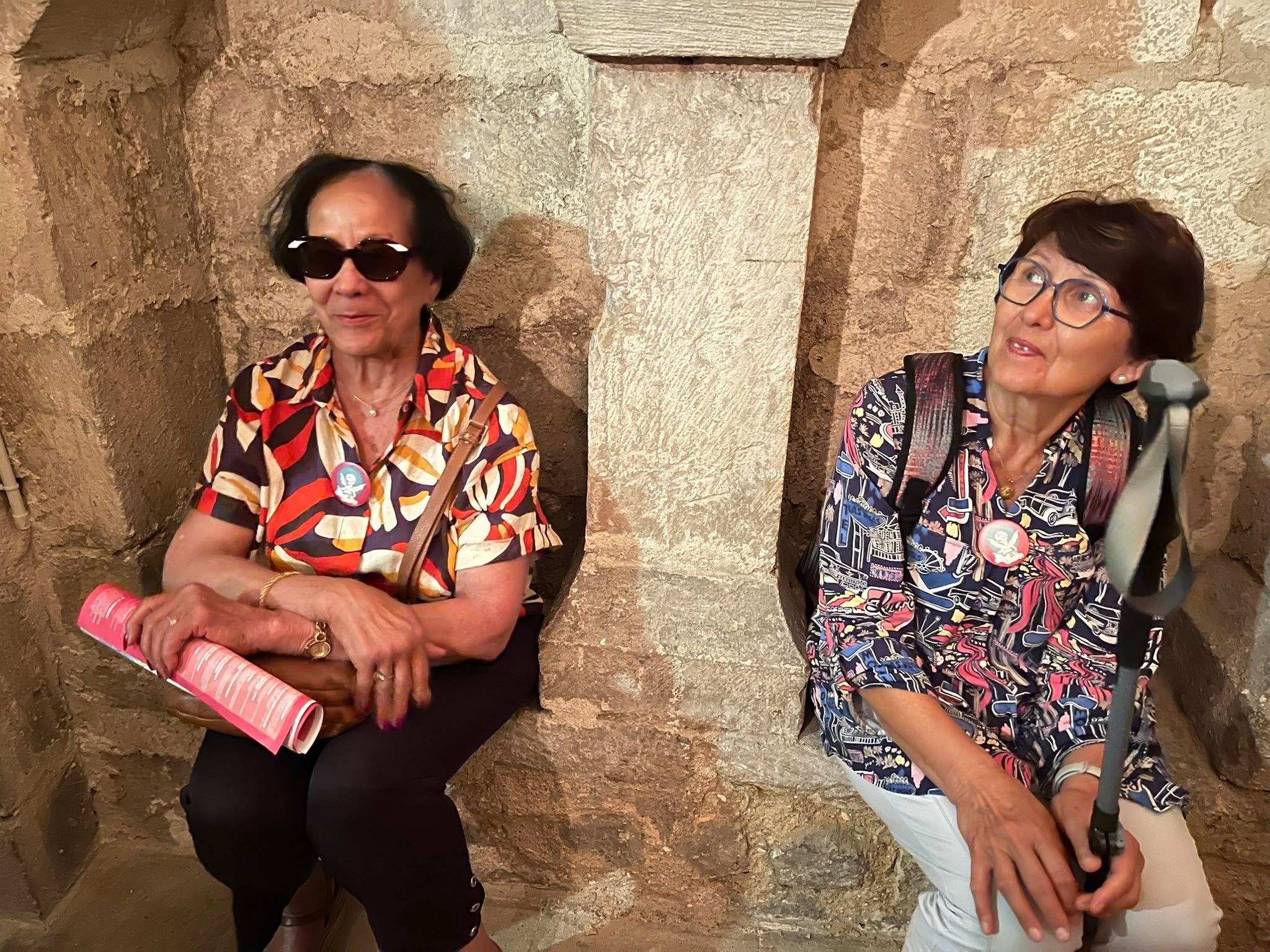 727445997_1305298581757965_6680857397769537608_n Two women sit against a rough stone wall, wearing colorful patterned shirts; one holds a pink rolled-up poster and wears sunglasses while the other watches to the side, wearing glasses.