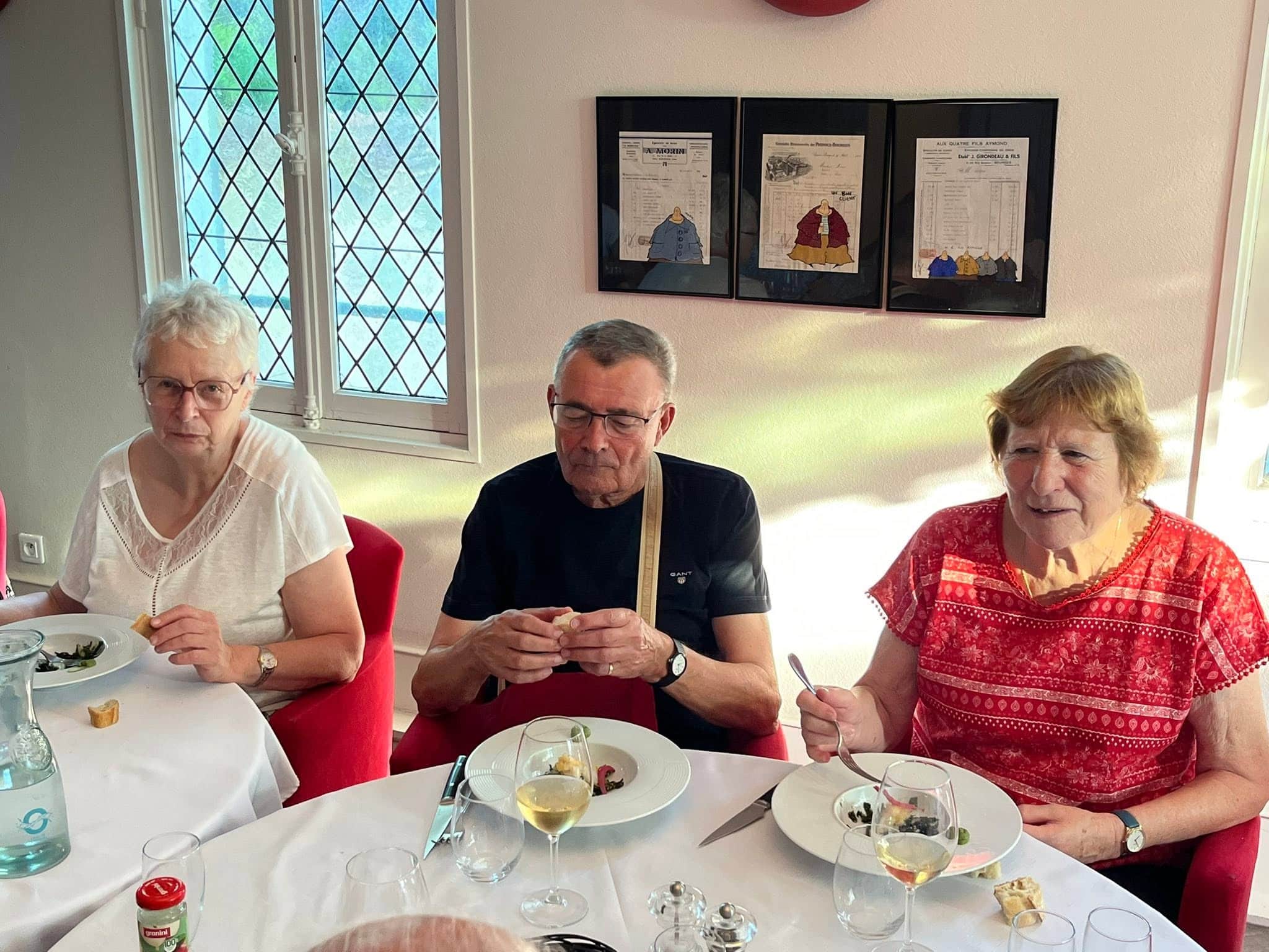 727519299_1305299921757831_4657899247843708573_n Three seniors sit at a table during a meal, with plates, glasses, and a bottle in front; background shows lattice windows and framed artwork on the wall.
