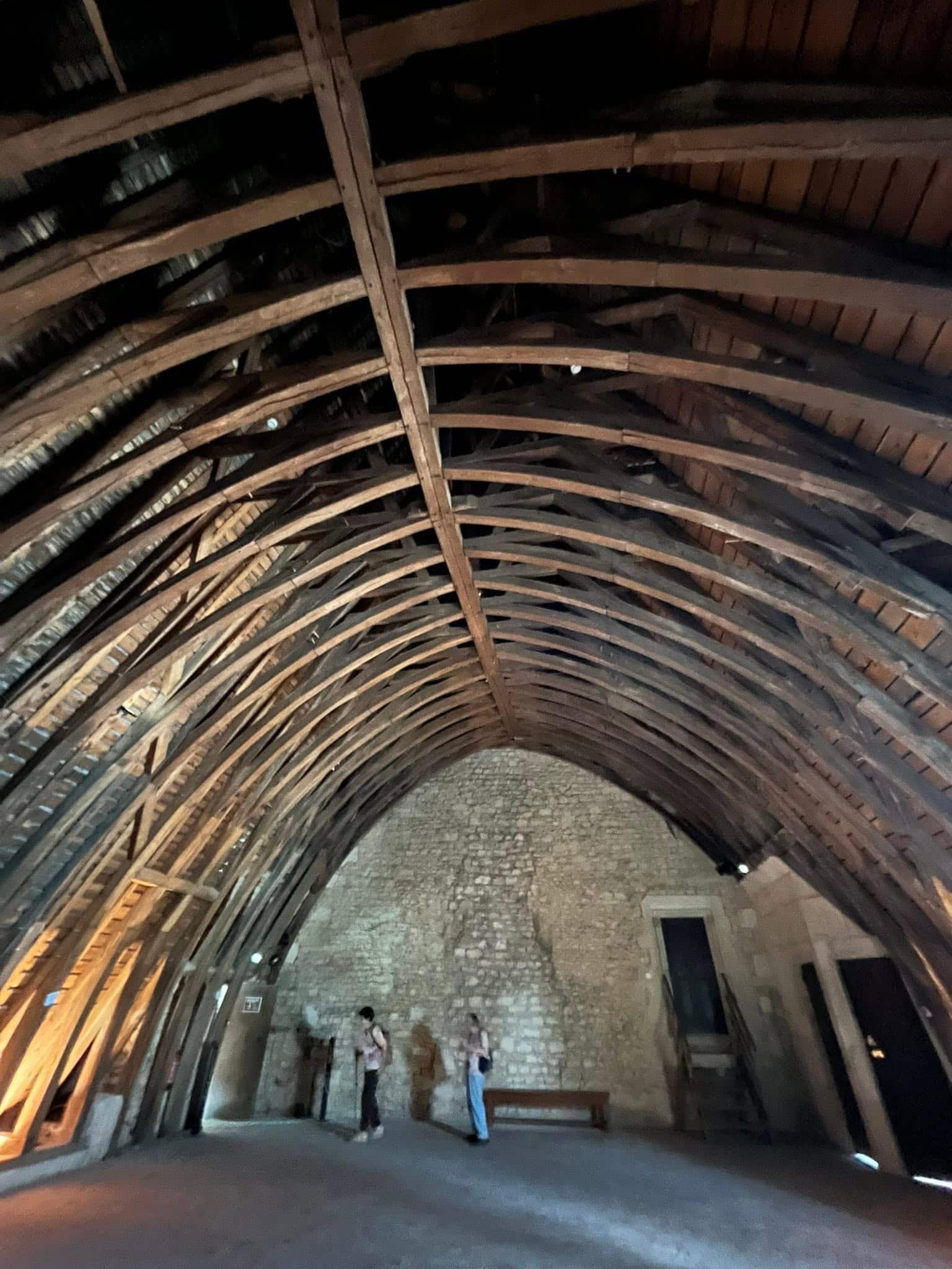 727519461_1306129371674886_1734452088929085184_n Interior of a large vaulted wooden roof with curved beams and stone walls, showing a few visitors inside a historic hall