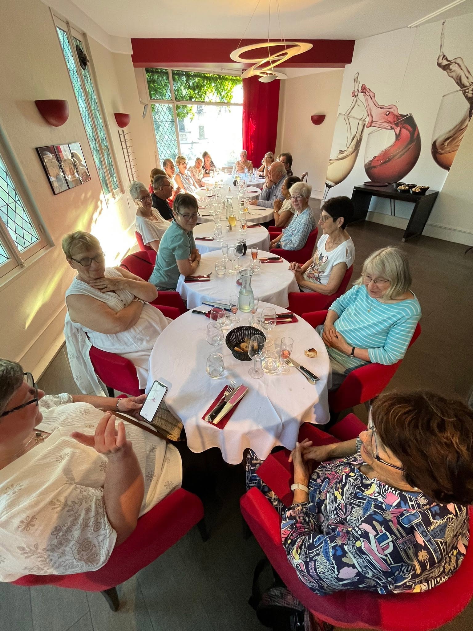 727542563_1305299588424531_903998233910232914_n Group of seniors gathered around a long table for a meal in a sunny dining room with red chairs and large wine mural on the wall.