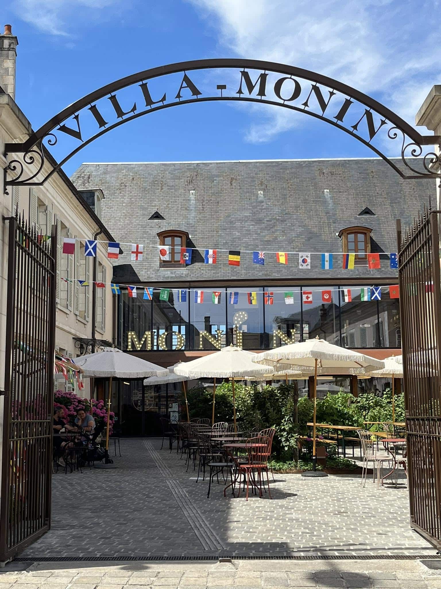 727658912_1305292161758607_3288447438177785276_n Archway with text 'VILLA MONNIN' over a courtyard cafe, flags strung overhead, and outdoor tables with umbrellas under a blue sky.
