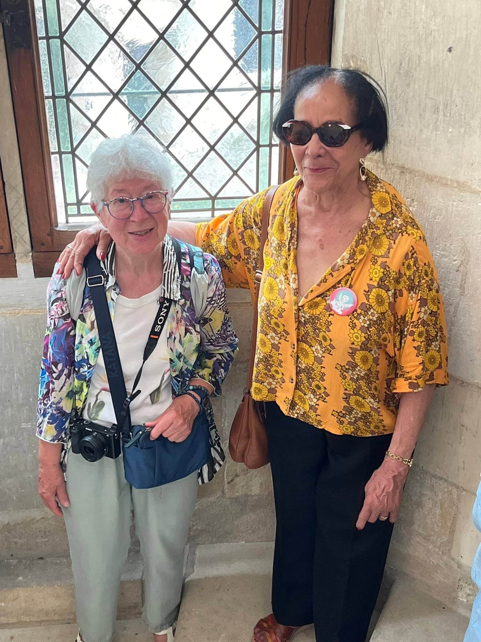 727715136_1306129501674873_7520336390038148359_n Two smiling women pose together indoors by a lattice window; the left woman has white hair, glasses, and a camera strap, the right wears sunglasses and a yellow floral blouse.