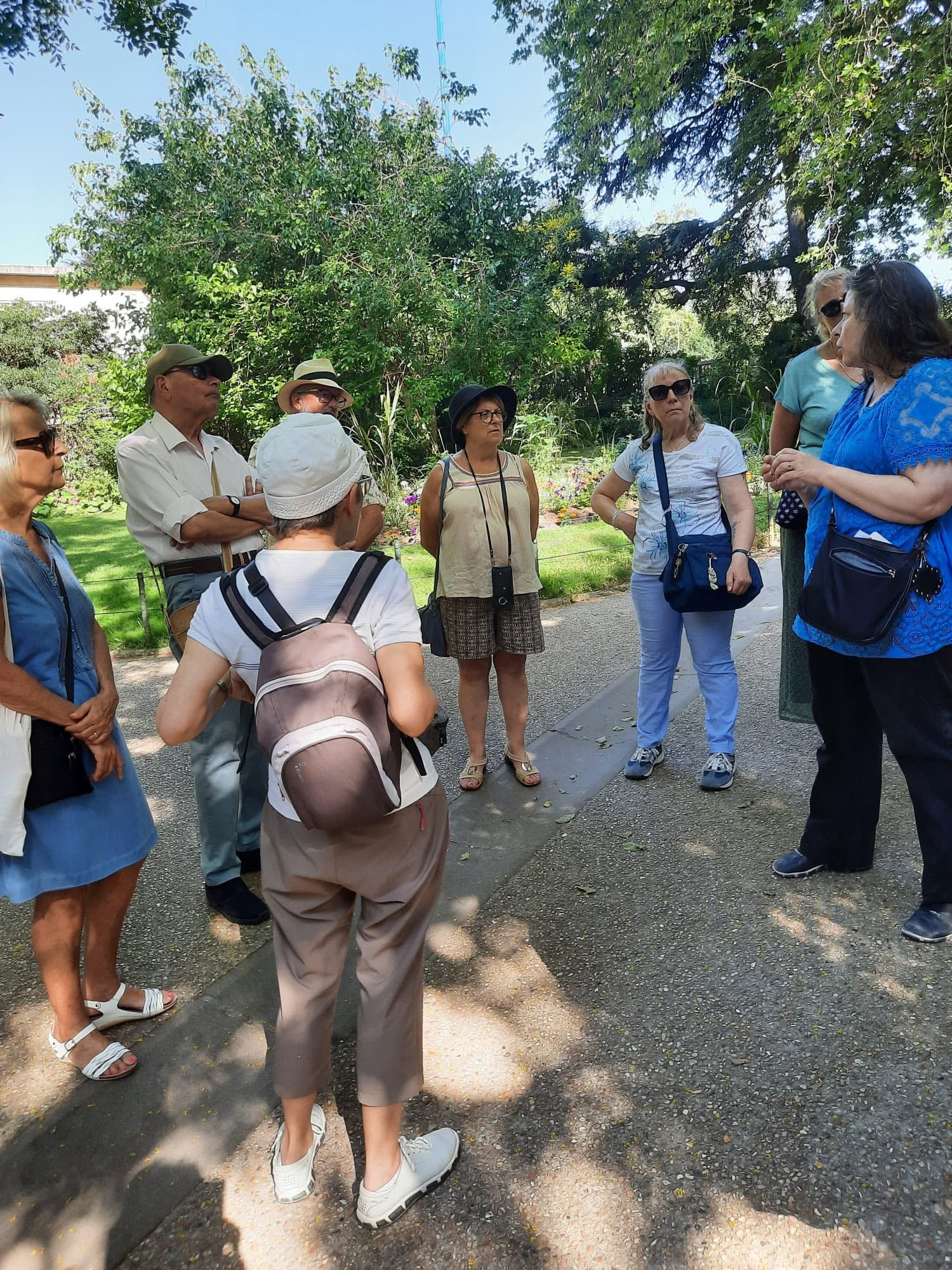 727884508_1308620154759141_2824656494579347006_n Group of adults standing in a circle and chatting on a sunny garden path, surrounded by trees and flowers.