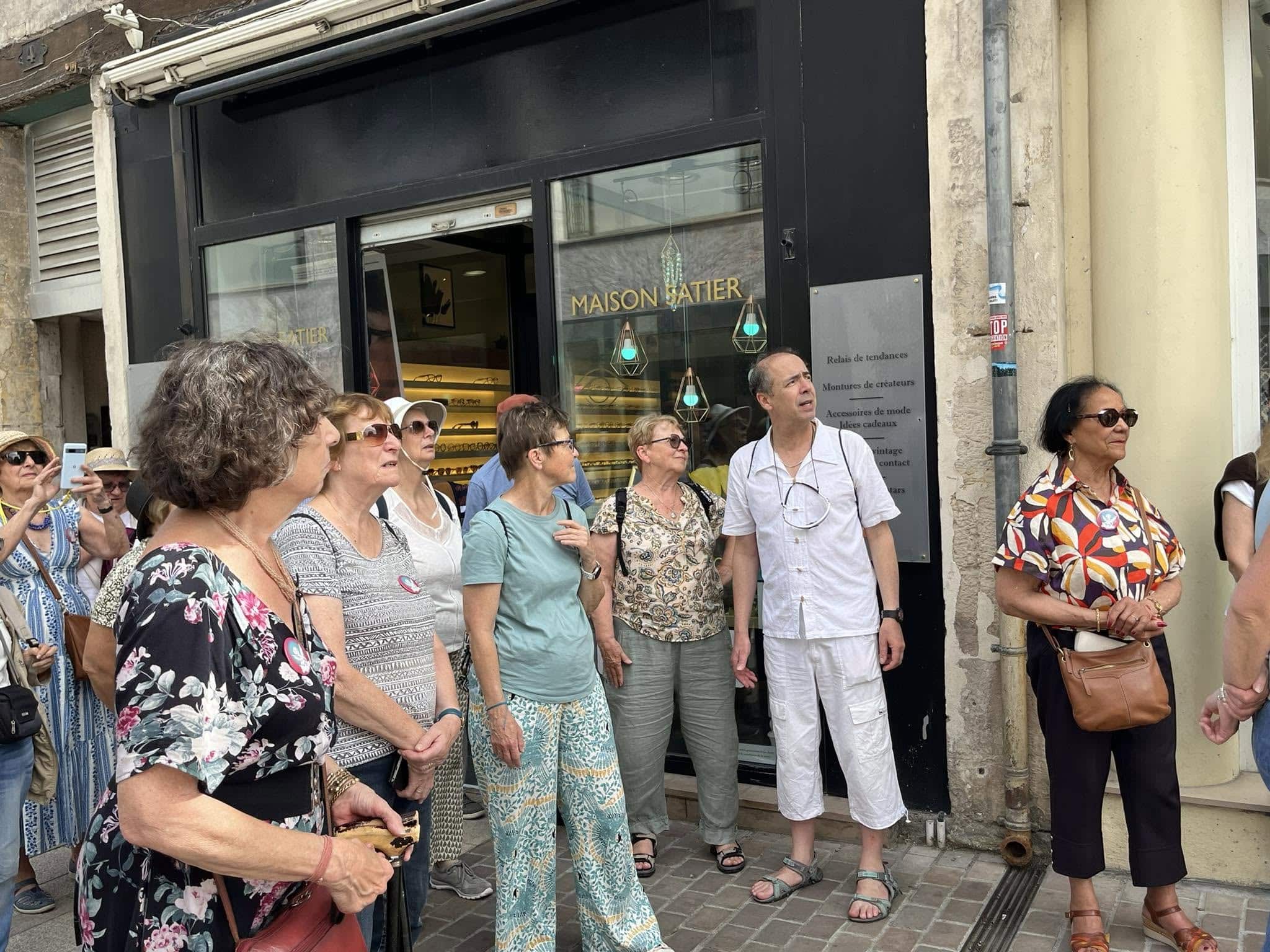 727919448_1305290831758740_255252506125729325_n Group of adults in summer clothing standing outside a shop window with the sign 'MAISON SATIER' visible behind them, on a city street.