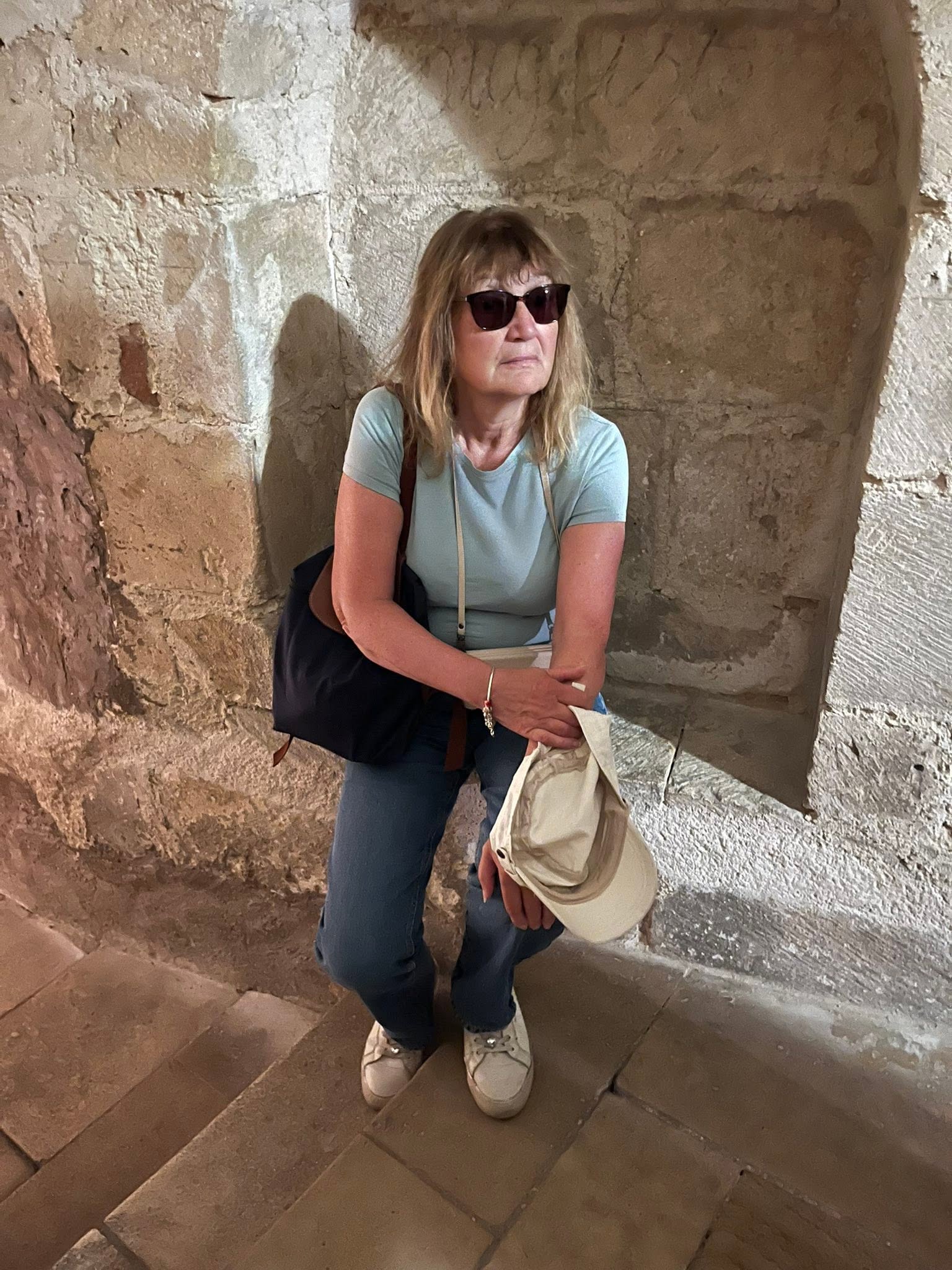 728234584_1305299151757908_5178429938092832039_n Woman wearing sunglasses and a light blue shirt stands on stone steps, holding a beige hat and a black bag.