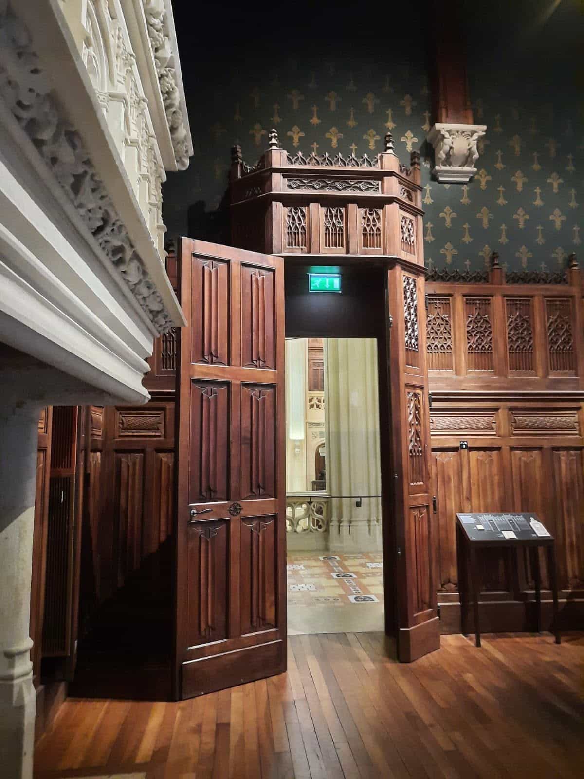 728254772_1310251217929368_976676439611570683_n Ornate wooden doorway and balcony with carved panels inside a historic room, dark wallpaper with fleur-de-lis pattern behind the door.