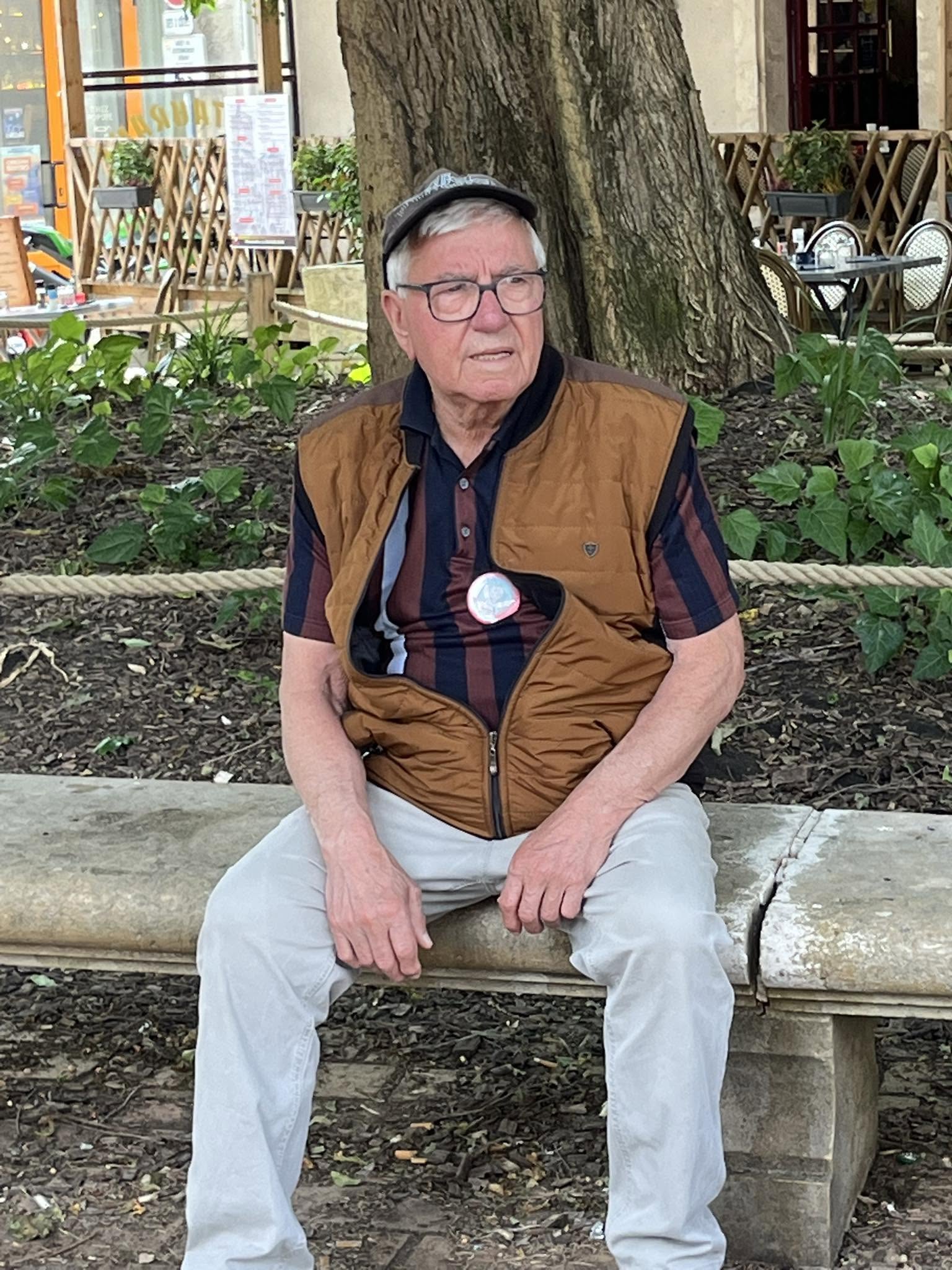 728294003_1305292221758601_2447341693170302948_n Older man with gray hair and glasses sits on a wooden bench outdoors, wearing a brown vest over a striped shirt, looking to the side with a large tree and cafe seating in the background.