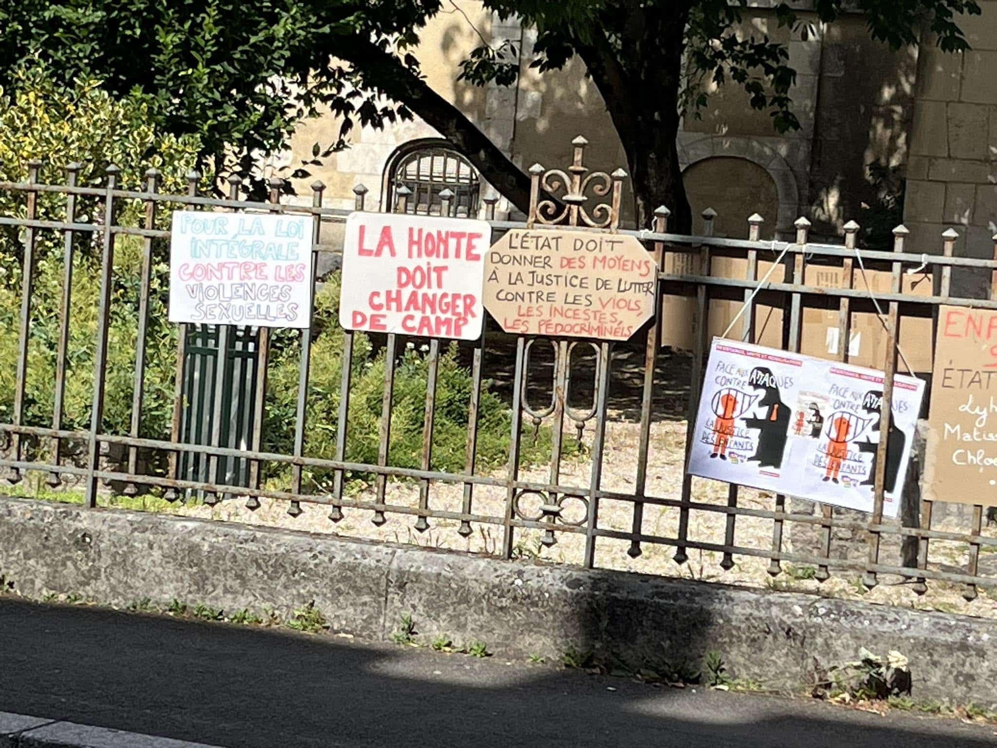 728427441_1305290885092068_9114889364427368410_n Protest signs attached to a metal fence, with messages in French about justice and ending violence against women, set against greenery and a stone wall behind the fence.