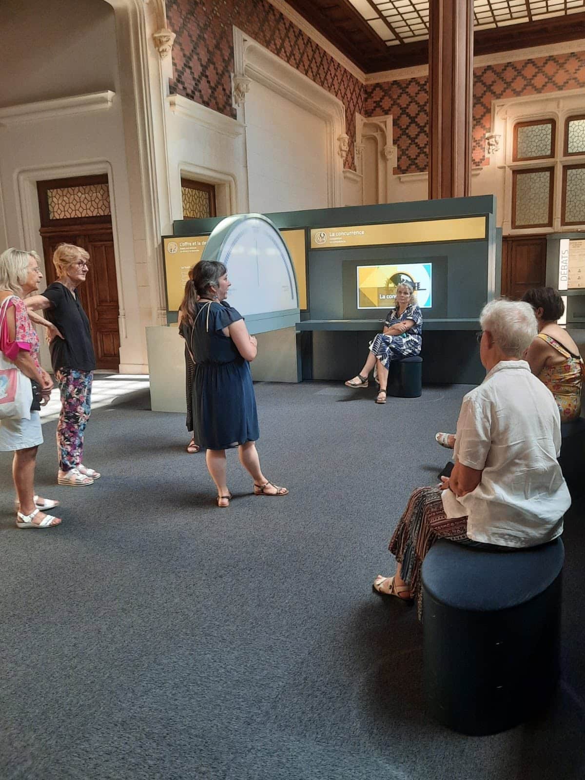 728893226_1310251407929349_345387480220374342_n Group of people listening to a woman presenting at a museum exhibit with a large circular display behind them. (Informative)