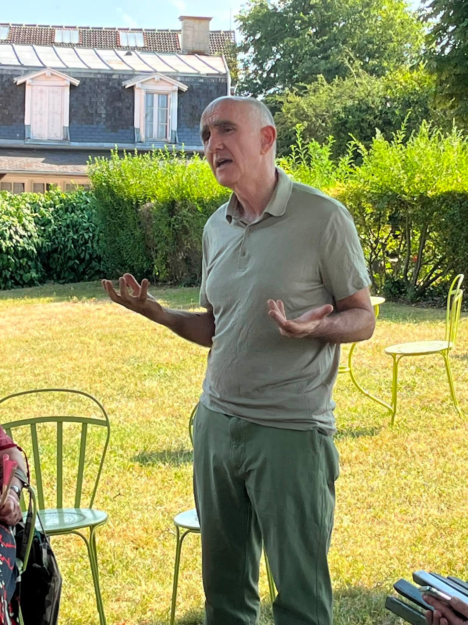 731059167_1311463994474757_111695270842894180_n Older man speaking outdoors, gesturing with hands in a sunny garden with green chairs and yellow grass behind him.