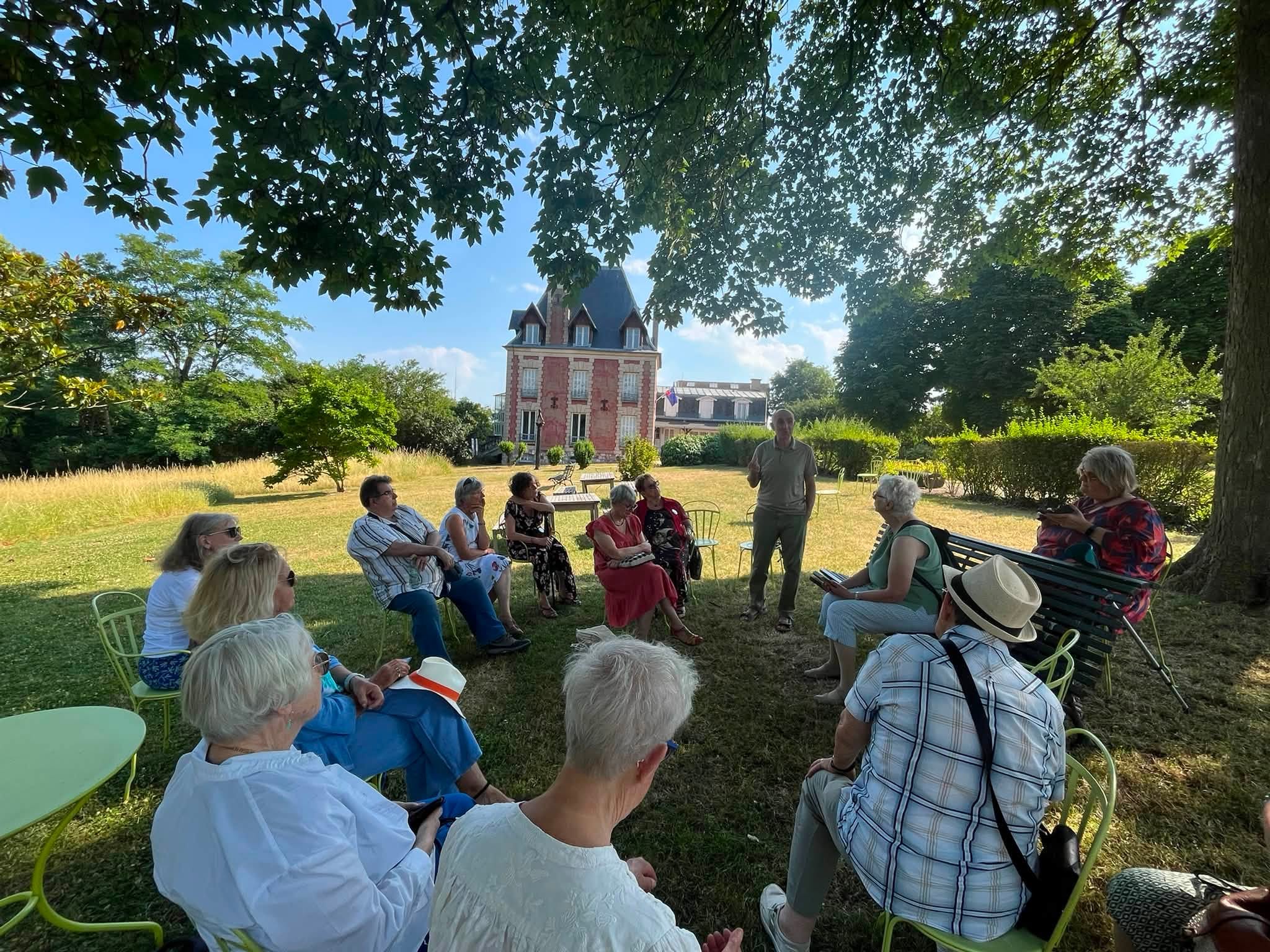 731068087_1311463854474771_6619405509989910702_n Group of seniors seated in a sunny garden, listening to a man speak, with a red brick building visible in the background.