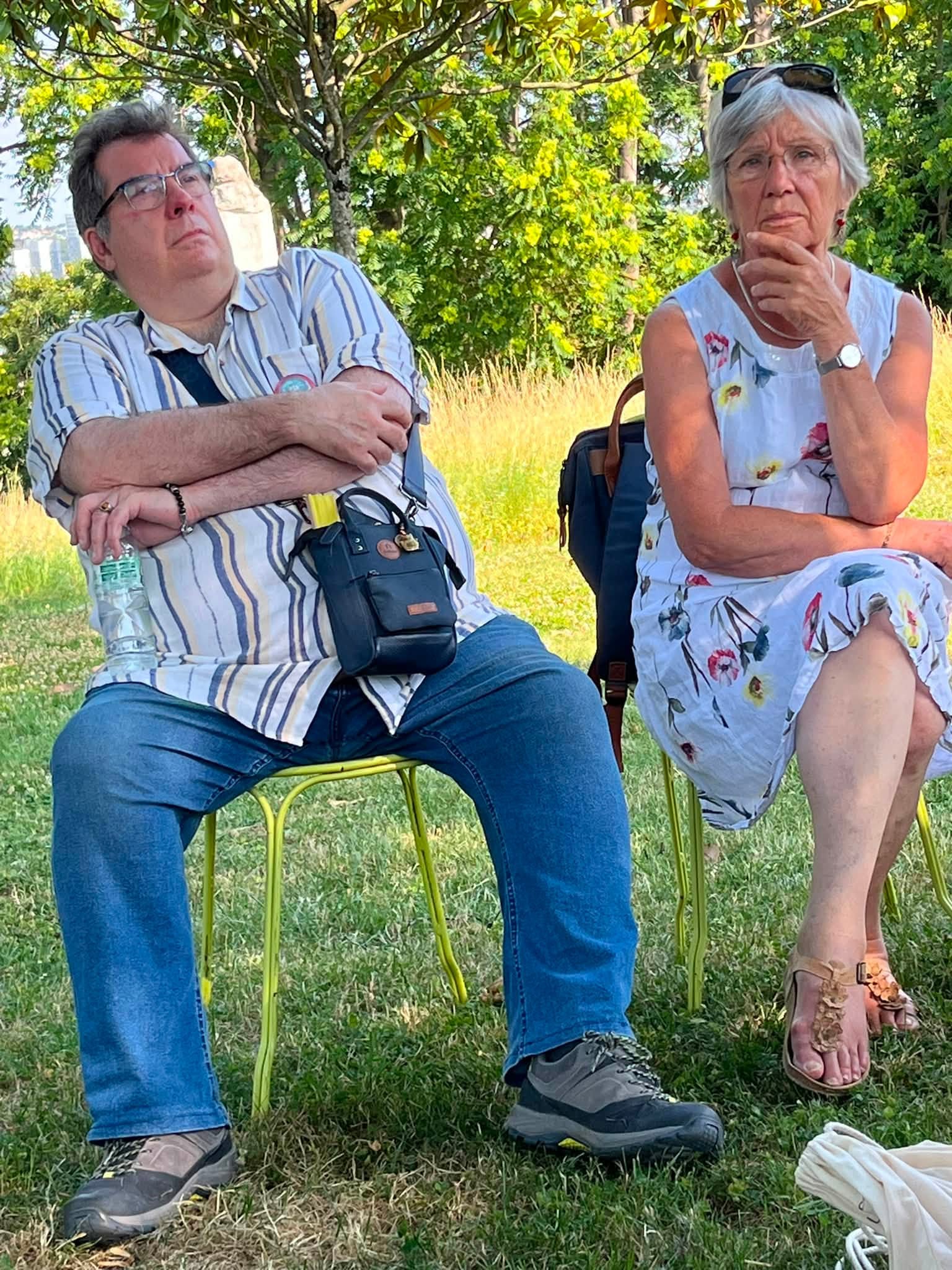 731131713_1311463884474768_2643724367783283699_n Two older adults seated on bright yellow metal chairs in a sunlit park, with a man on the left crossing his arms and a woman on the right touching her chin.