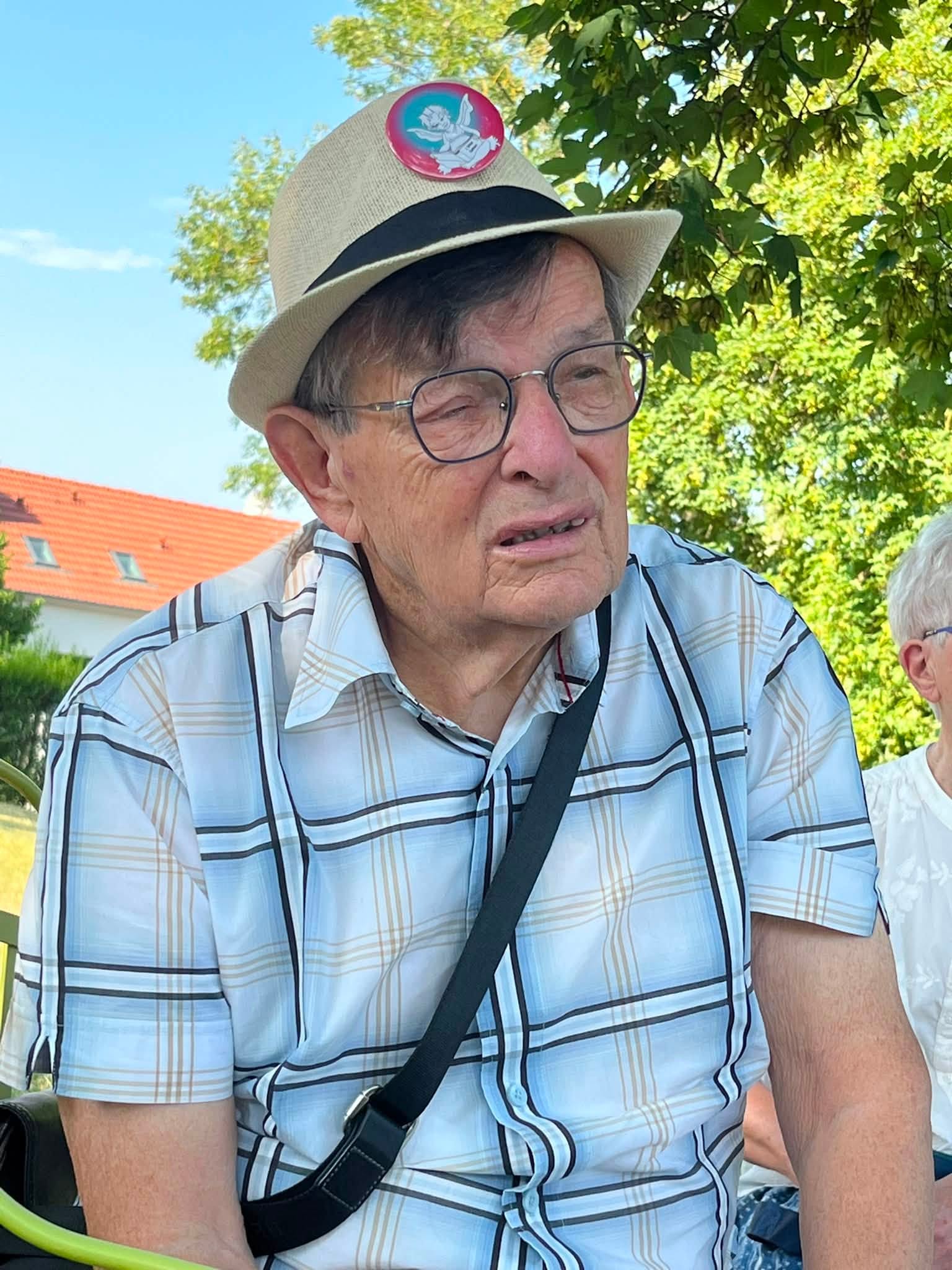 731131726_1311463831141440_6561116543208916998_n Older man wearing a beige fedora, glasses, and a light plaid shirt sits outdoors with trees in the background.