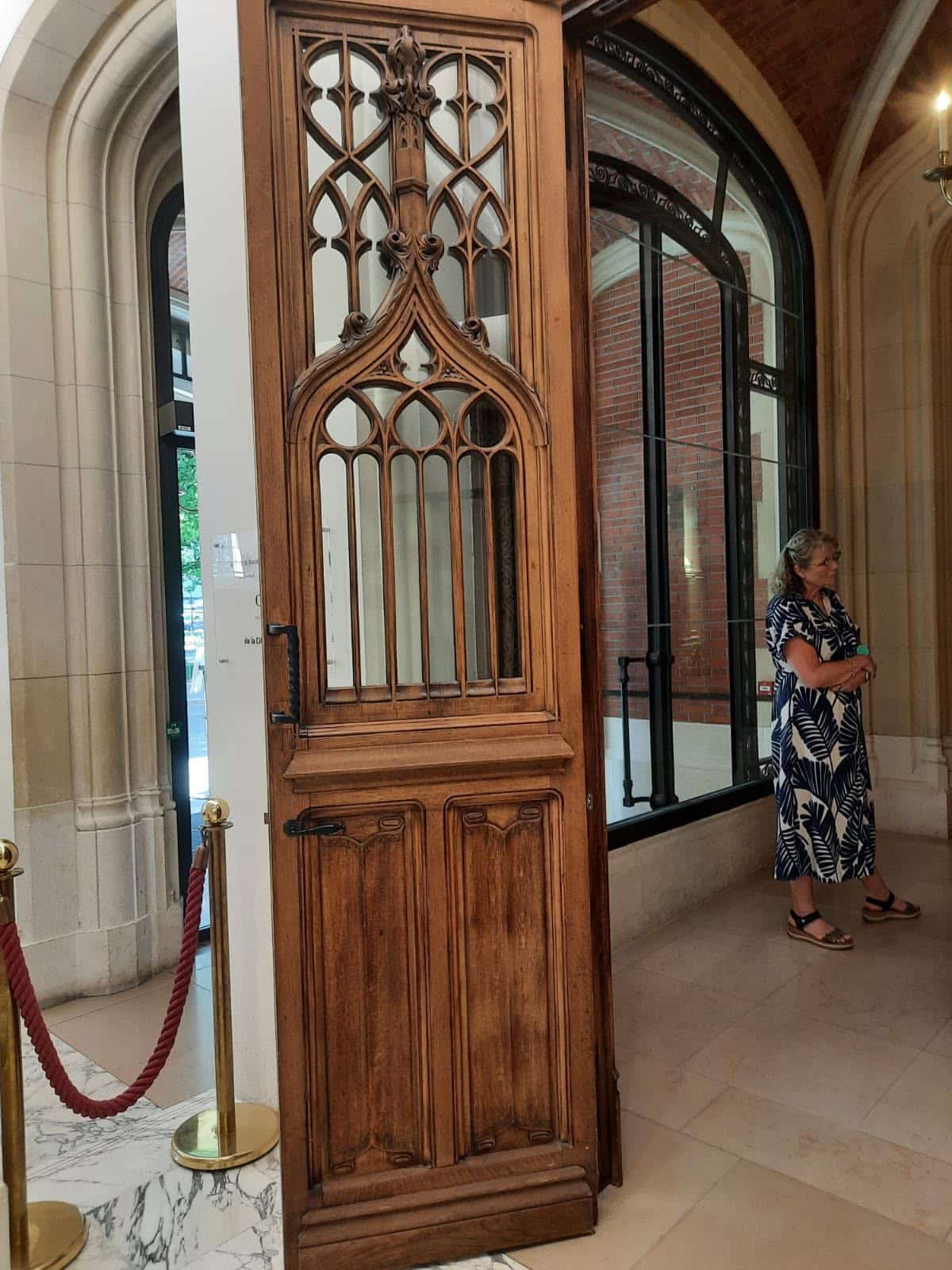 731138122_1310251174596039_4788697821273277284_n Tall carved wooden door with Gothic lattice panels in a museum hall; a woman in a blue patterned dress stands nearby by a large glass wall.