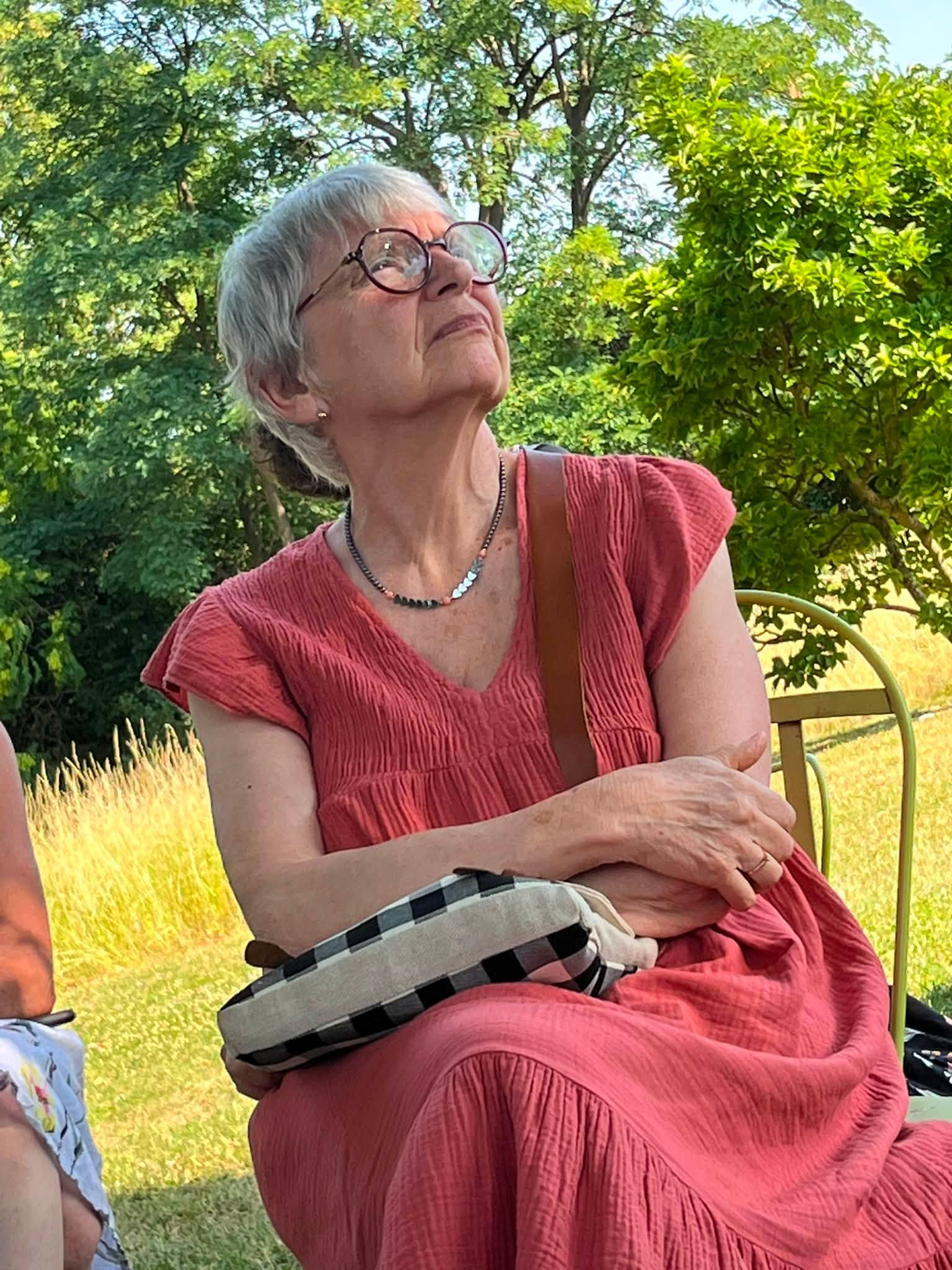 731173263_1311464107808079_5052856441311577106_n Older woman with short gray hair and round glasses, wearing a red dress, seated outdoors among greenery and trees.