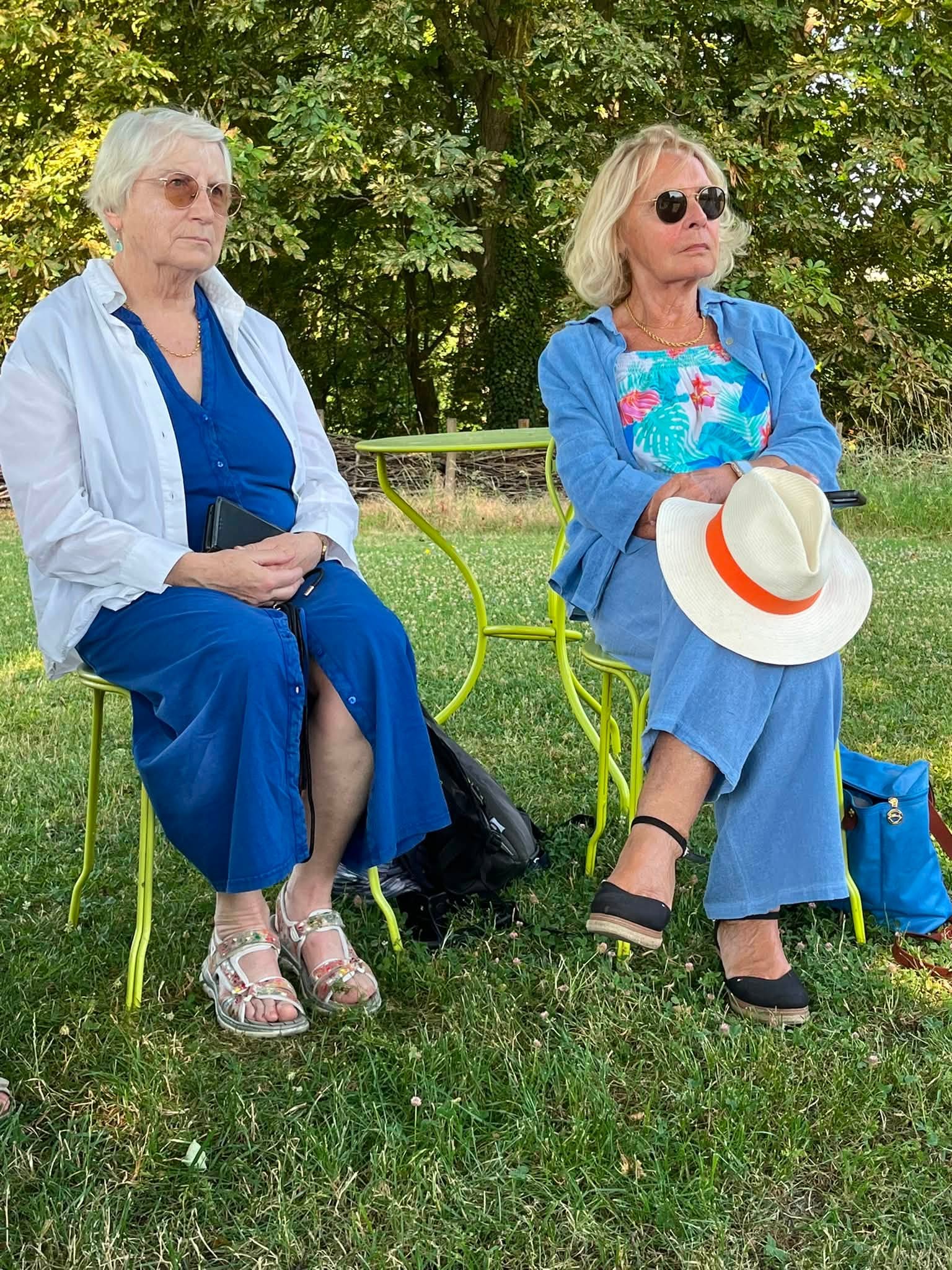 731232921_1311464074474749_6567696017161822409_n Two elderly women sit on bright green metal chairs in a grassy park, wearing blue outfits and sunglasses; one holds a hat in her lap.