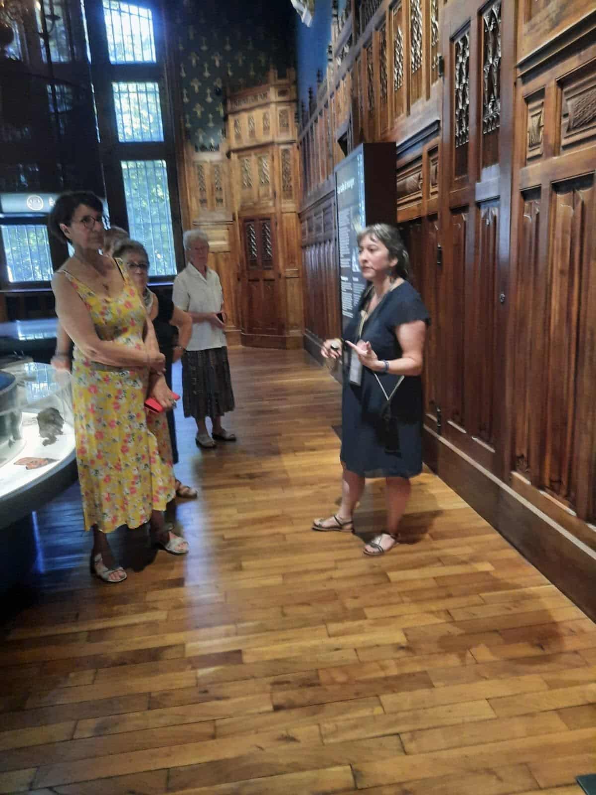 731898365_1310251377929352_2095907604221354033_n Group of four visitors listening to a guide in a blue dress inside a museum with ornate wooden paneling and a glass display case at left.