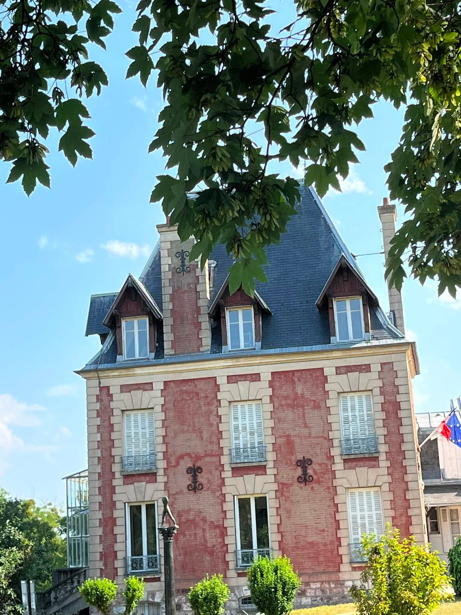 733427730_1311463817808108_2726452626802639627_n Red brick townhouse with a slate roof and dormer windows, stone trim, and a flag on the right, framed by leafy trees against a blue sky.