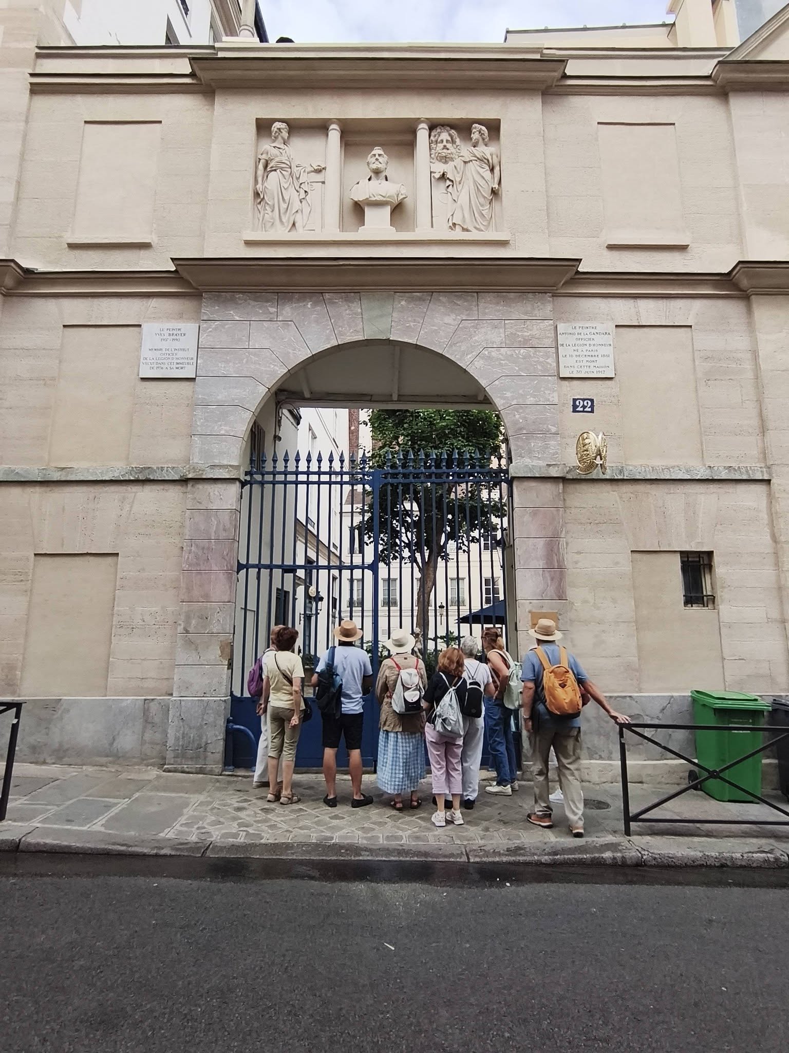 735636552_27010033035363545_4984861457658490323_n Tour group with backpacks gathered at a stone arch entrance of a historic building, blue gate, with plaques on either side and statues above the arch.