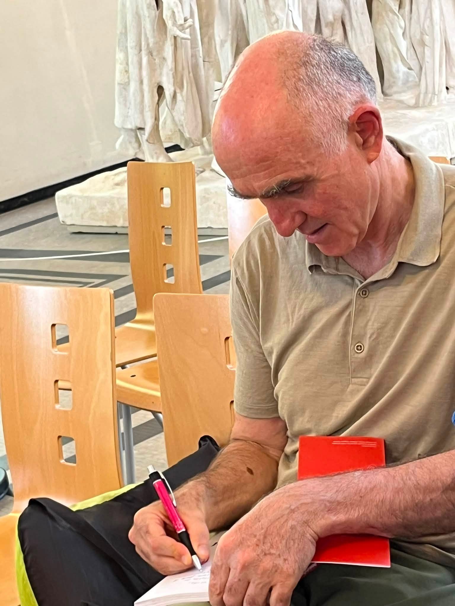 741882267_1322793436675146_3635653952174110641_n Older man seated and writing on a notepad with a pink pen, surrounded by wooden chairs in a classroom-like setting.