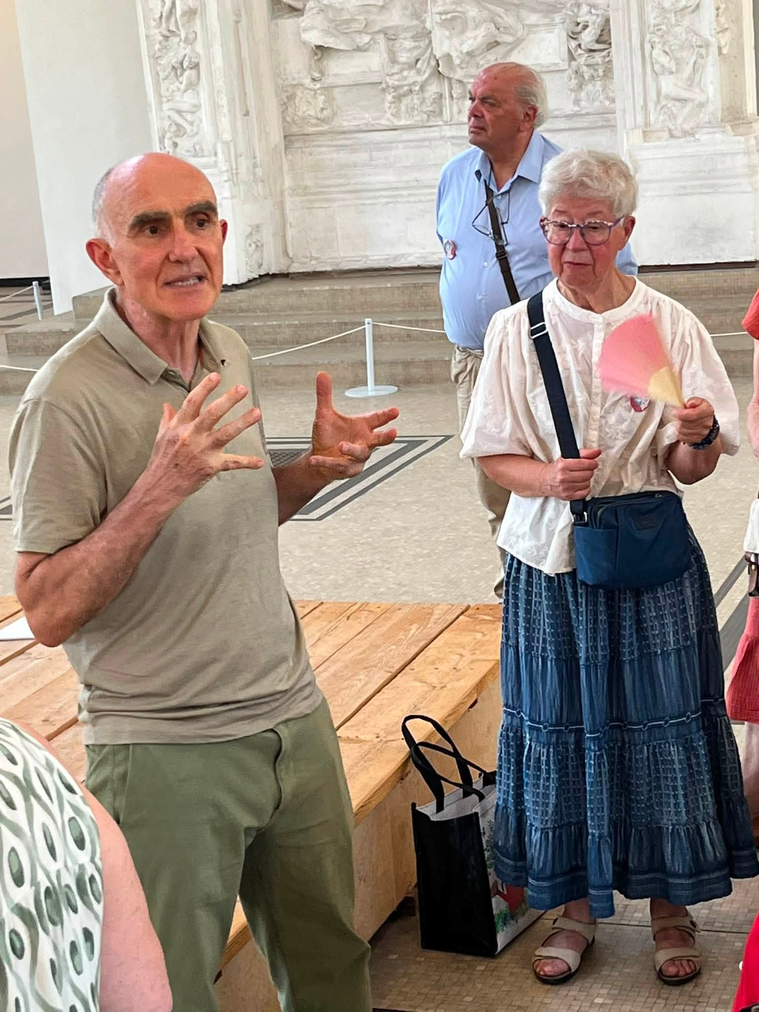 741948246_1322793063341850_6205592256805450883_n Three seniors in a museum gallery: a man in a beige polo gesturing with his hands while to his right a woman in a white blouse holds a pink fan, marble relief wall in the background.