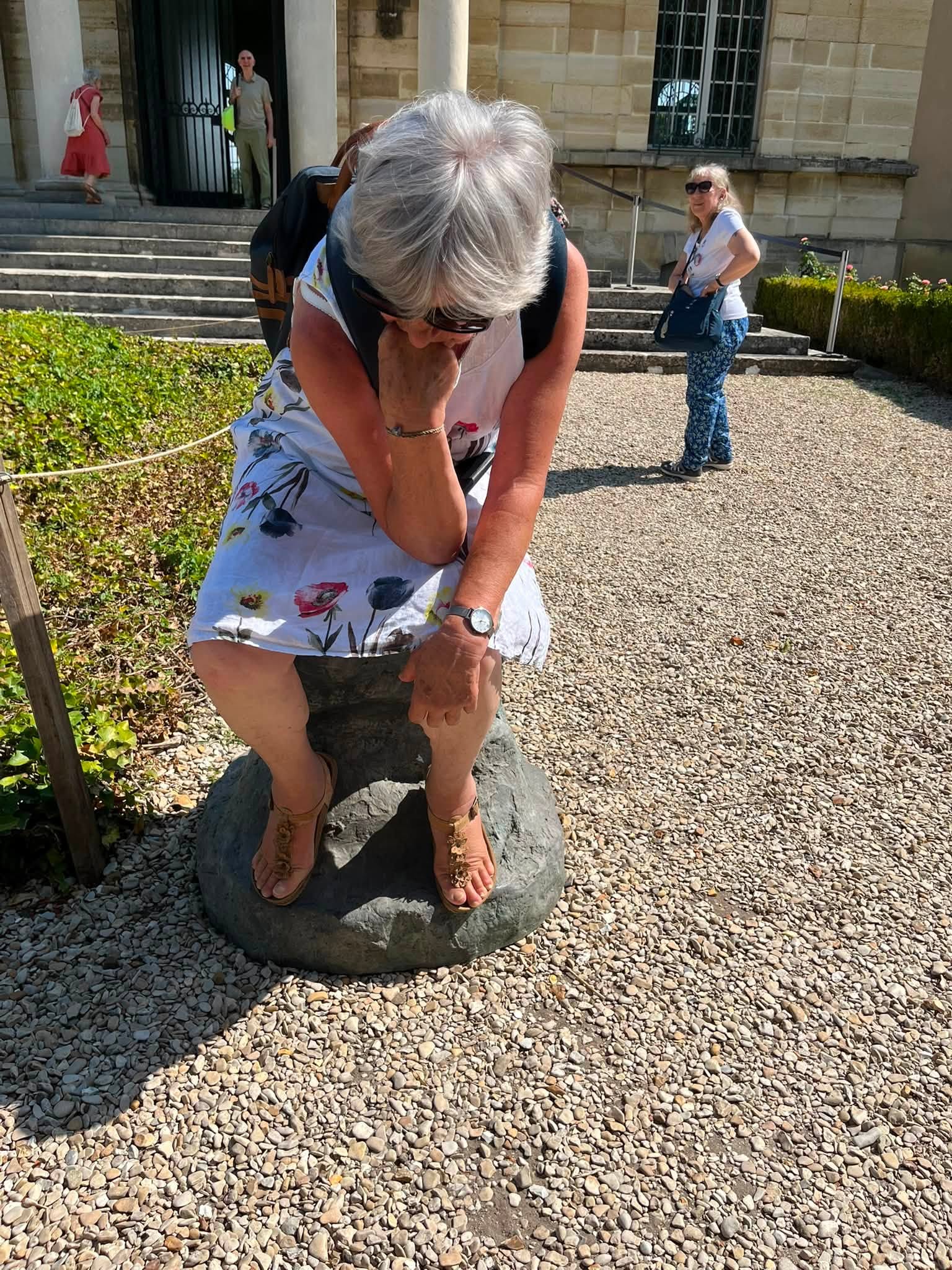 742059623_1322793410008482_5104386565089551087_n Older woman with white hair bending over a rock statue outdoors, wearing a floral dress and sandals with henna on her feet.