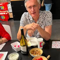 742145271_1320691093552047_4124250082777232816_n Man in a patterned shirt sits at a table with a bottle of white wine and bowls of snacks, looking at the camera.