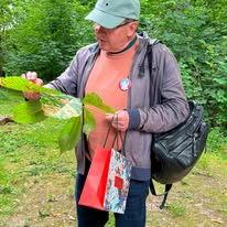 742208374_1320691080218715_8953023117415740953_n Man in a cap and jacket in a forest, inspecting a large leaf while carrying a backpack and a red gift bag.