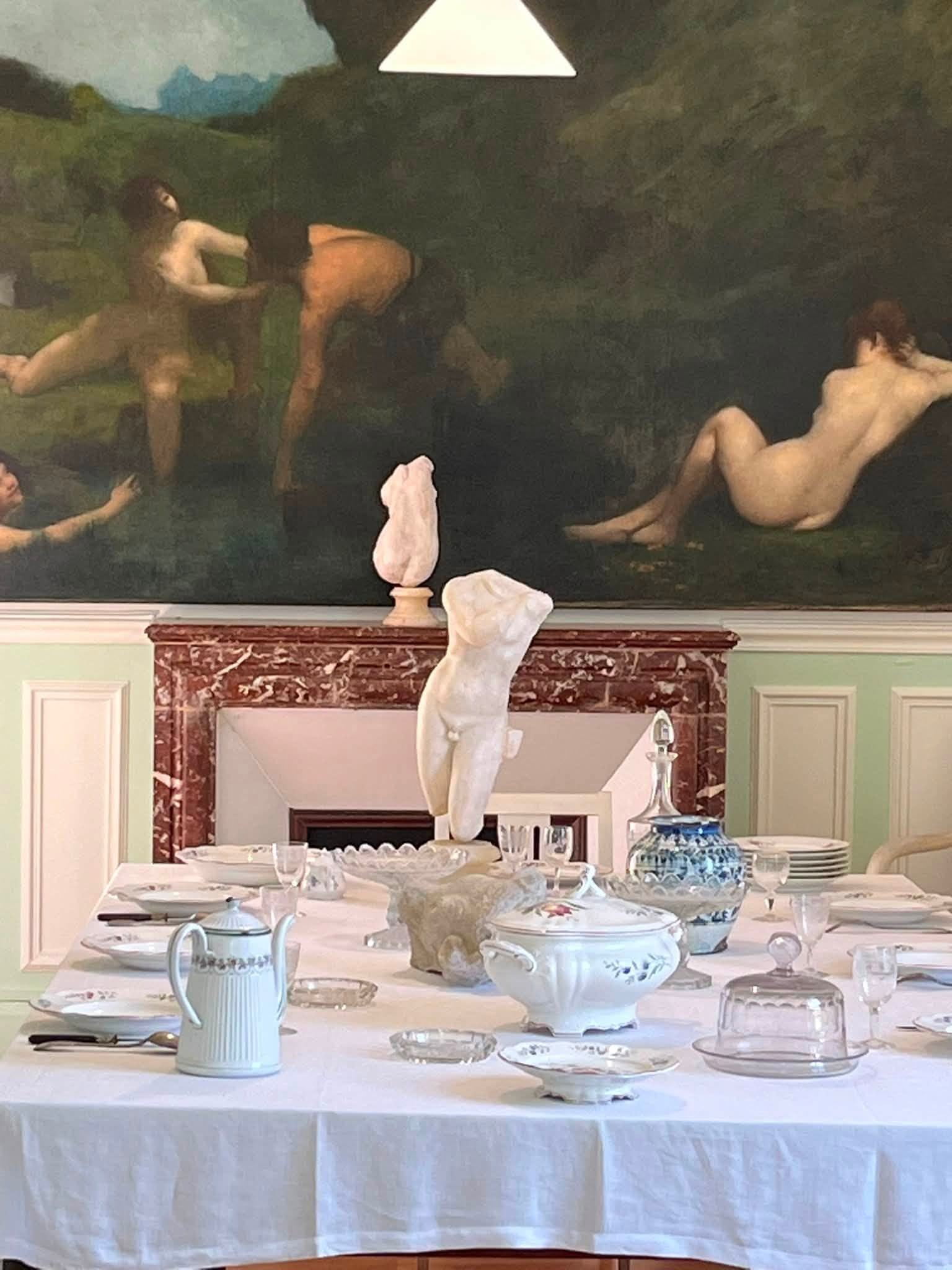 742308972_1322793170008506_7679979502930724062_n Formal dining table set with fine china, glassware, and a pale blue teapot on a white tablecloth, in a room with green paneling and a landscape painting behind them.