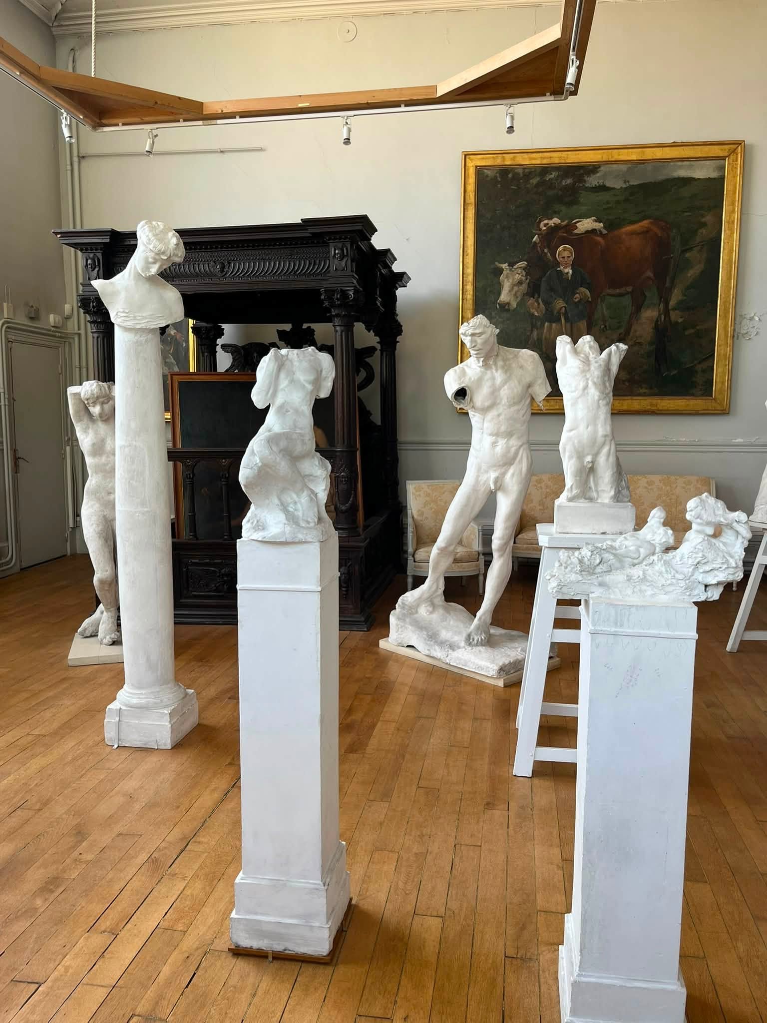 742786763_1322793086675181_739917876317107097_n White plaster sculptures and busts on white pedestals in a gallery, with a dark ornate cabinet and a gold-framed painting on the wall behind.