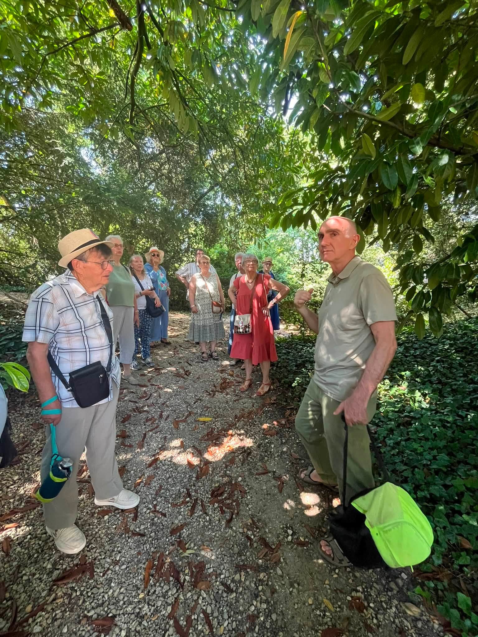 742876278_1322793073341849_4981729121053962145_n Group of elderly people on a gravel path shaded by trees, listening as they tour a garden.