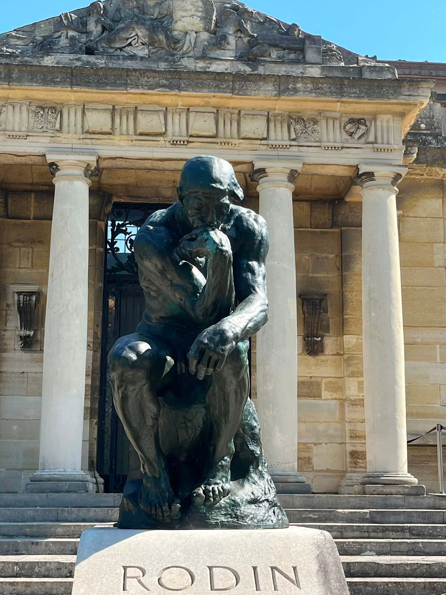 743203736_1322793456675144_2484722709764734784_n Bronze seated statue of a thinker in front of a colonnaded historic building; base reads RODIN.