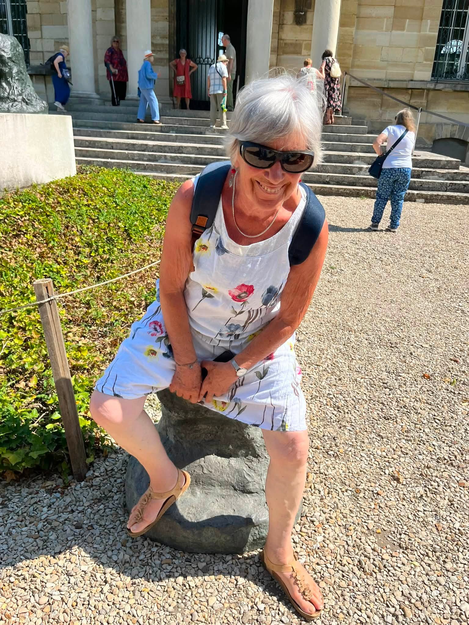 743975354_1322793300008493_2329500459892205970_n Older woman with white hair and sunglasses smiling, wearing a white floral dress and brown sandals, leaning on a stone post outside a building.