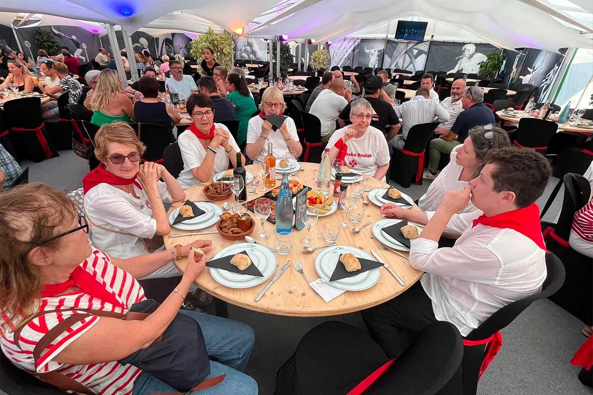 Group of people sitting around a round wooden table at a festive banquet under a white tent, wearing white tops with red scarves, enjoying bread and drinks together.