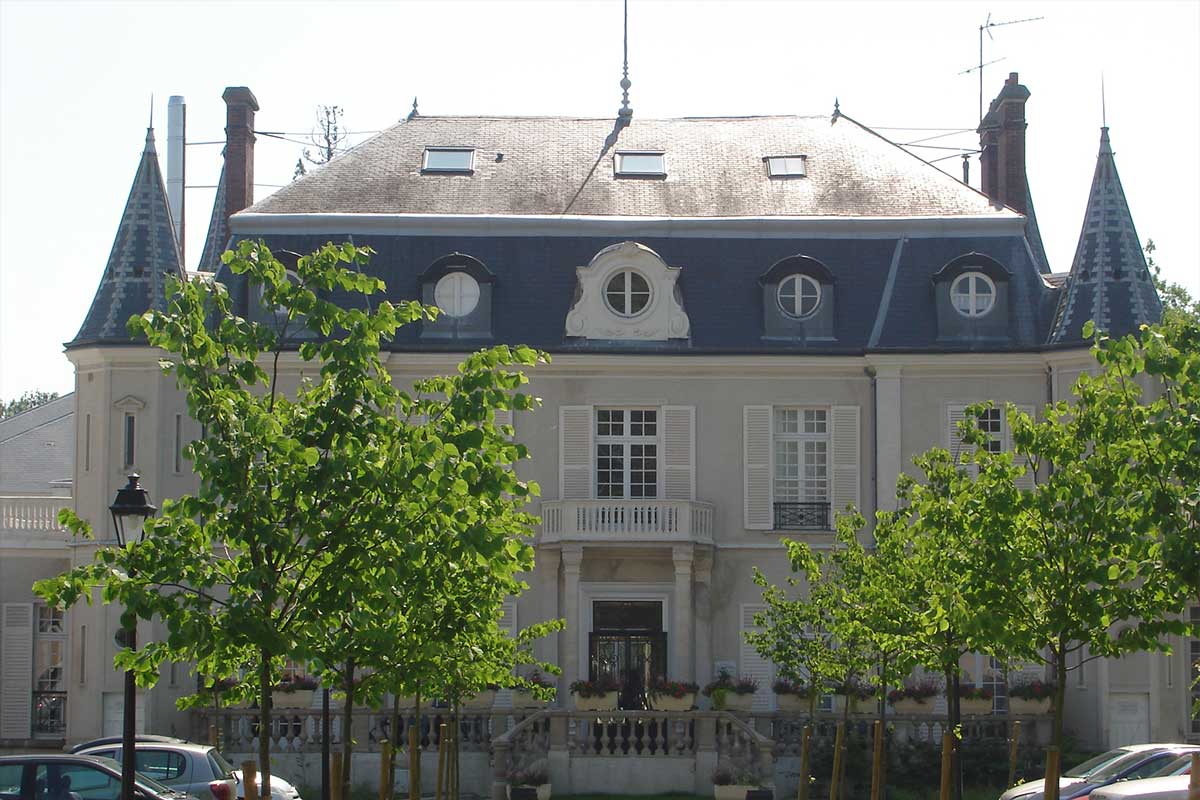 Grand mansion with a blue slate roof, ornate dormer windows, and a stone balcony, framed by green trees in front.