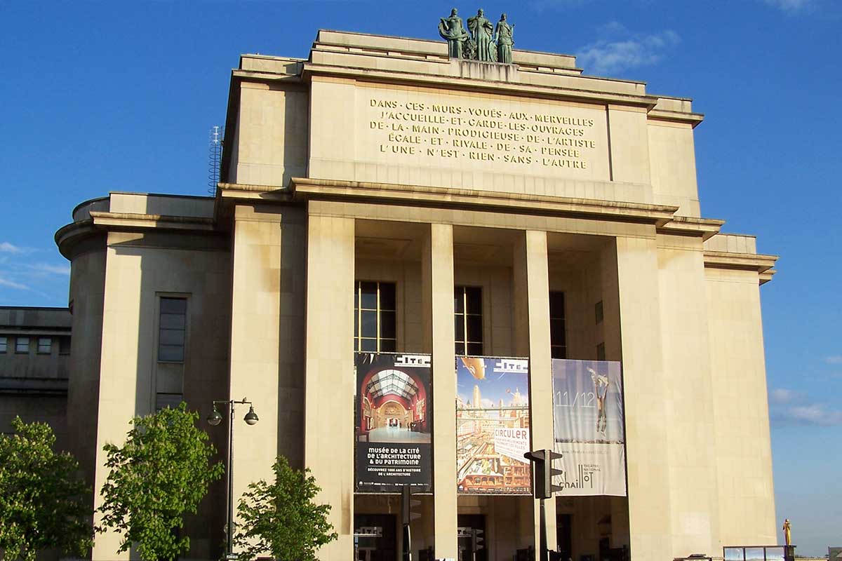 Beige neoclassical museum facade with tall columns, statues on top, and three banners hanging between the pillars against a blue sky.